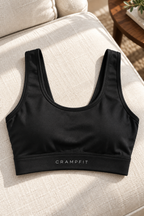 Black CRAMPFIT cropped U-neck sports bra on linen, showing the neckline, wide underbust band, logo and fabric texture.