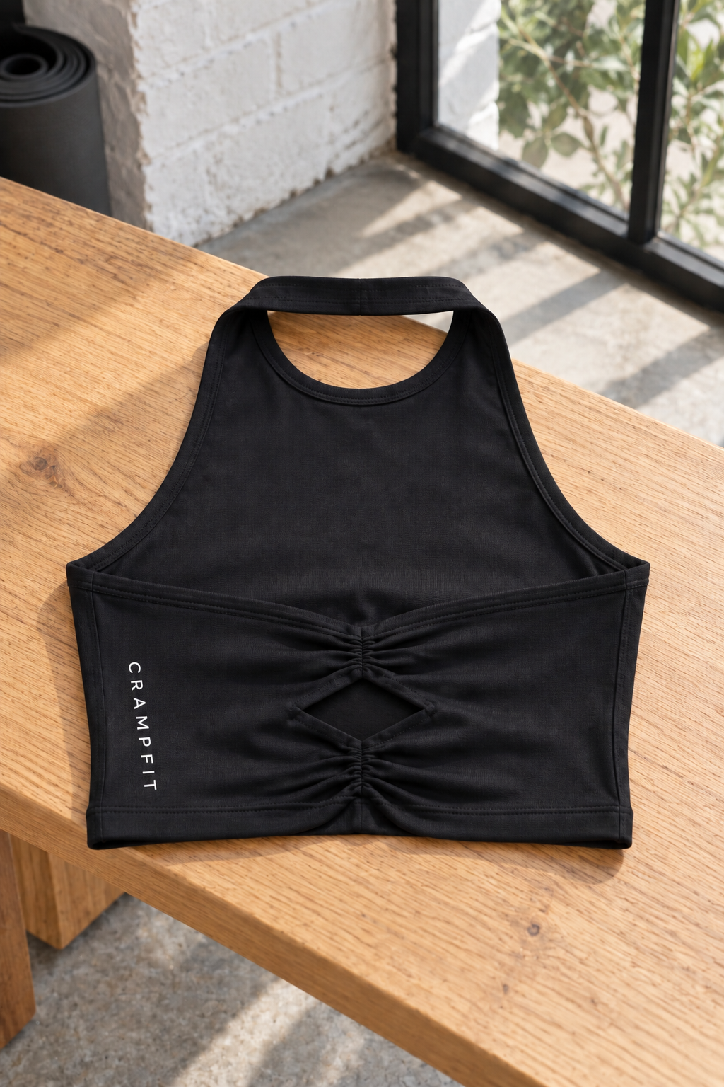 Black CRAMPFIT halter yoga sports bra laid on an oak bench, showing back ruching, diamond-shaped opening, neck strap and fabric texture.
