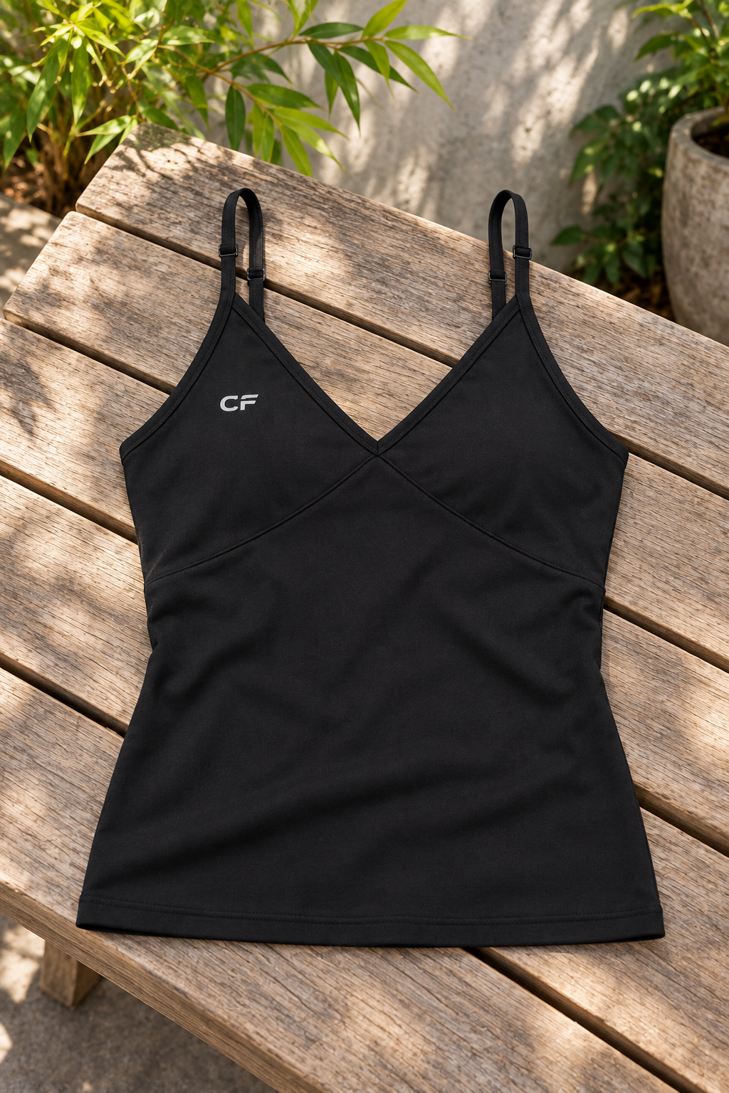 Black CRAMPFIT V-neck yoga tank laid on a wooden bench, showing its adjustable straps, shaped front seams, CF logo and fabric texture.