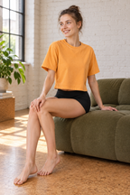 Model seated in CRAMPFIT orange crop tee with black athletic shorts in a bright loft.